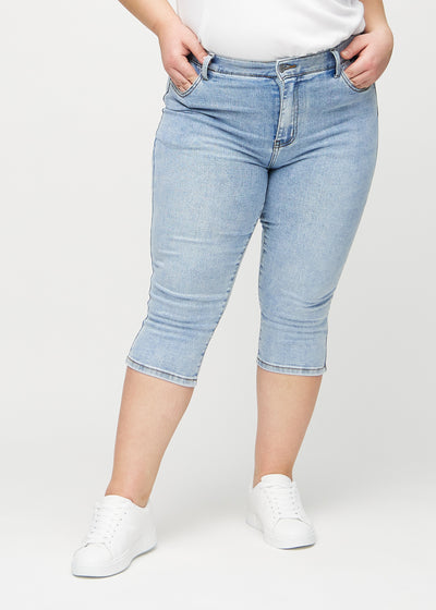 Lys denim skinny capris, modelnavn Waves, som sidder tæt til benet og slutter lige under knæet på en plus-size model, set forfra.